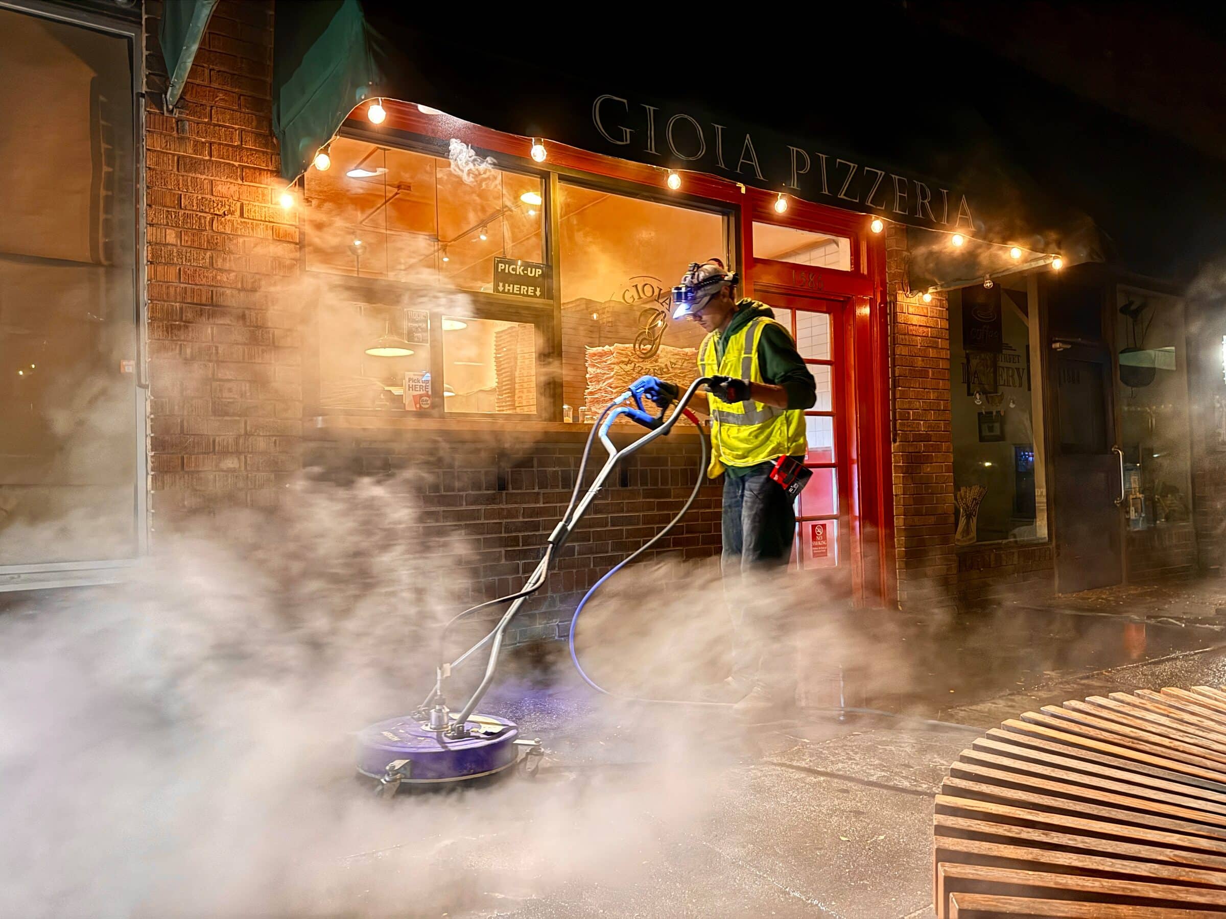 A technician in a reflective vest and headlamp runs a hot-water surface cleaner over a restaurant's front sidewalk at night as steam rises off the concrete.