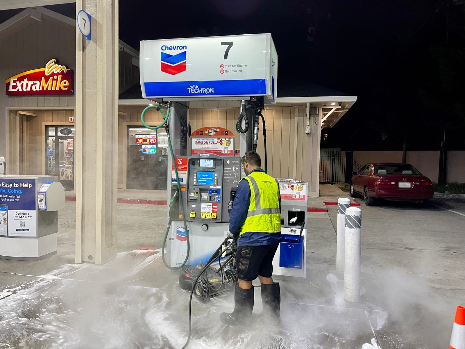 A technician in a reflective vest scrubs the concrete pad at a fuel pump at night as hot water and foam spread across the island.