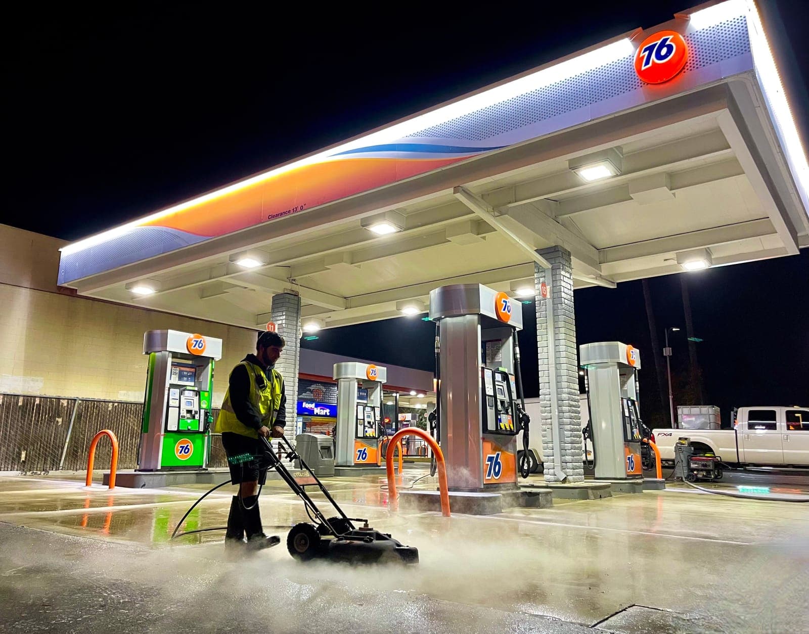 A Bay Area Exterior Cleaning technician runs a hot-water surface cleaner across a gas station forecourt at night, with steam rising off the concrete beside the fuel pumps.