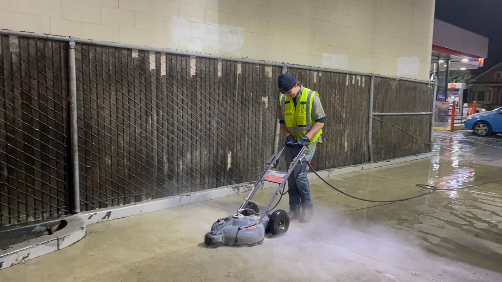 A technician runs a surface cleaner along the fence line at the edge of a gas station lot at night, leaving clean concrete behind.
